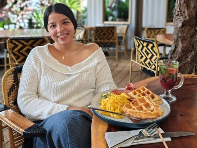 MarelingRossi, mit leicht geöffnetem, weißem Pullover, der ihre Brüste bedeckt, lächelt und sitzt an einem Holztisch mit Waffeln, Eiern und einem Getränk, und trägt blaue Jeans..