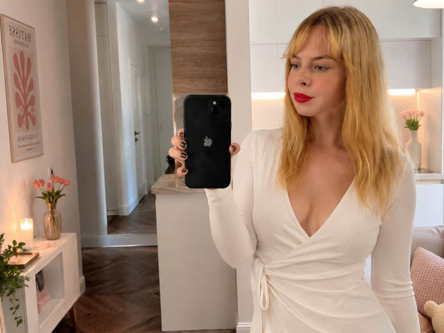 Nellie Vanduyne, con i capelli biondi, il rossetto rosso, un abito bianco a V profondo, che tiene in mano un iPhone nero, mostra il seno, in piedi in una stanza moderna, con pavimento in legno, pareti bianche e candele..