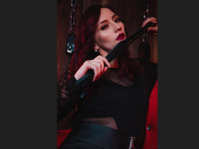 Aysha Smith, con cabello rojo, sosteniendo un látigo de cuero negro, vestida con una blusa negra transparente y una falda de cuero, con una expresión seductora, labial rojo oscuro, fondo de madera, iluminación roja..