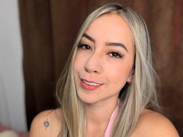 EmilyBonnett with blonde hair, small nose ring, light makeup, pink halter top, smiling, brown eyes, freckles, small tattoo on left shoulder, and a cute, sexy face.