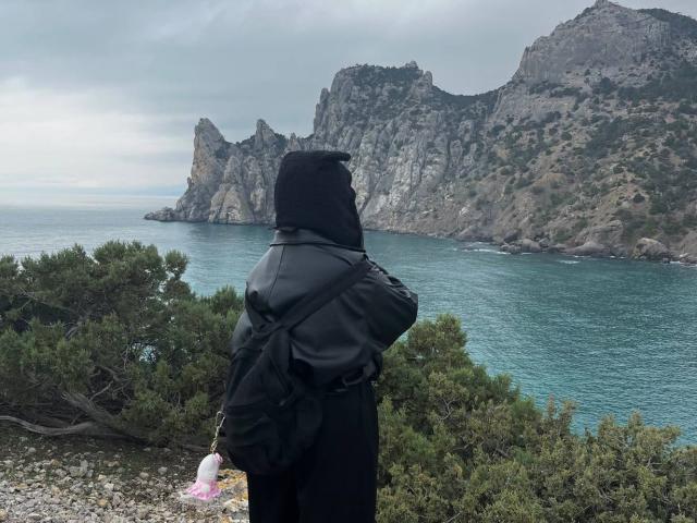 LynnaMitton est debout, dos à la caméra, portant un sweat à capuche noir et un sac à dos, et regarde une côte rocheuse et une mer bleue. Ses fesses sont légèrement visibles..