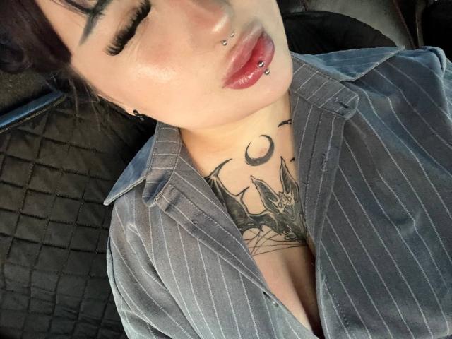 Lynna Mitton, con dei piercing al naso e alle labbra, indossa una camicia a righe e mostra un tatuaggio di pipistrelli e una mezzaluna sul petto, con gli occhi chiusi..