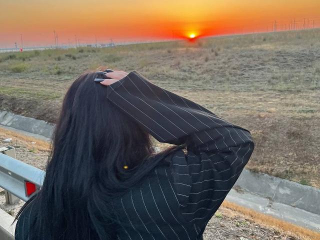 Lynna Mitton, con lunghi capelli neri, indossa una camicia nera a righe sottili e si copre il viso con la mano, in piedi a lato di una strada durante il tramonto..