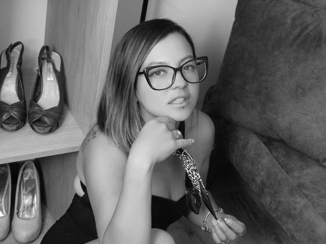 ArianaBlonde with glasses, holding a tie, has her hand near her chin, showing her tits in a black dress, sitting on a couch, with shoes in the background.