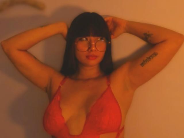 Genesiss with red lace bra, glasses, and black hair, arms raised, showing tattoos on arms, medium tits, and red lipstick, in warm orange light.