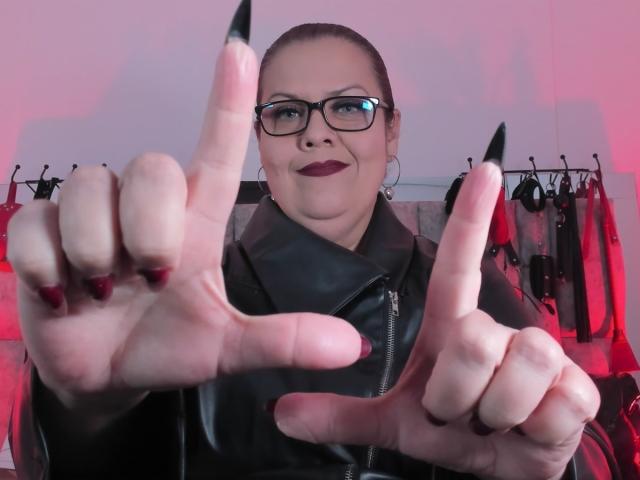 VanasMoka met een bril, donkere lipstick en een zwarte leren jas, die lange, puntige nagels aan beide handen laat zien, met wijzende vingers, lachend, in een kamer met rode verlichting..