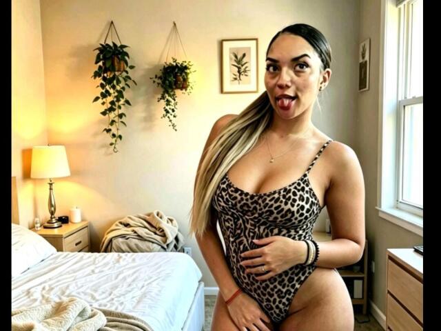 BrianaPratt, avec une longue queue de cheval blonde, la langue tirée, portant une combinaison une pièce à motifs léopard, montrant ses grandes seins, dans une chambre avec un lit, une lampe et des plantes suspendues..