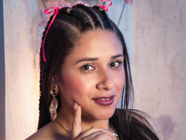 EmmyRossi, avec ses tresses foncées, ses rubans roses et ses boucles d'oreilles ornées, touchant son menton avec un doigt, souriant, montrant ses lèvres rosées et ses yeux marrons..