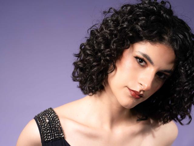 RoxyGraff has curly black hair, dark eyes, and red lipstick. She's wearing a black, studded top. Her tits are small and she's looking at the camera.