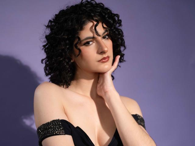 RoxyGraff, con i capelli neri ricci, la pelle chiara e un'espressione sensuale, che si tocca la guancia, indossa un abito nero con spalline scoperte e borchie argentate, su uno sfondo viola..
