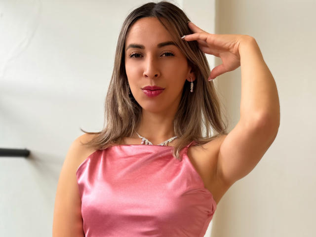 NikkyCranford met bruin haar, een roze satijnen jurk en diamanten oorbellen, die haar rechterarm omhoog steekt, haar oksel laat zien en een lichte frons heeft. Borsten zijn zichtbaar onder de jurk..