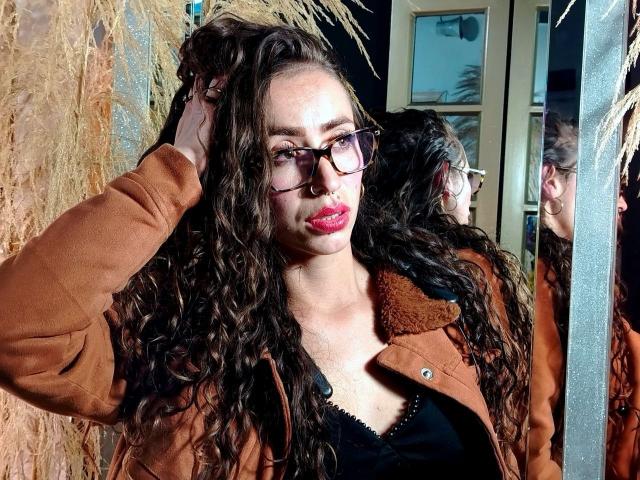 NatashaKraucer with long curly brown hair, glasses, red lipstick, brown coat, black top, holding hair, mirror reflection, dark background, night, slightly serious expression, natural light.