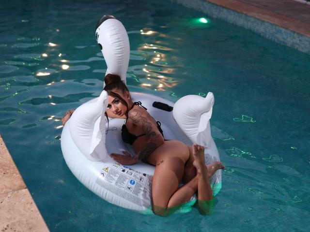 KayaBelle is lying on a white inflatable swan in a pool, wearing a black bikini, showing her tattooed arm and butt, with green water reflecting on her skin.