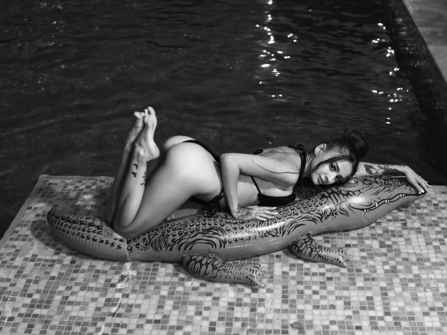 Black-and-white photo of KayaBelle lying on a crocodile inflatable, wearing a black thong bikini, with tattoos on her thigh and ankle, in a pool. Her ass is raised.
