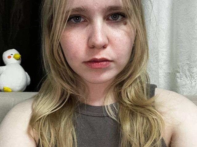 TiffaniMacandog hat helle Haut, blaue Augen und blondes Haar. Sie trägt ein graues, geripptes Tanktop und auf der linken Seite befindet sich ein kleines, weißes Entenspielzeug. Ihr Gesicht zeigt leichte Rötungen..