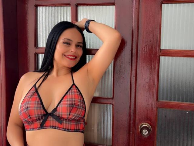 CharlotteCrofort with black hair, red plaid bra, black bow, smiling, arm raised, red doors, white glass panels, red wristband, red lipstick, curvy, medium tits, cute.