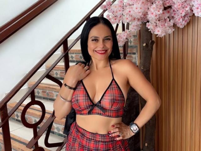 CharlotteCrofort with black hair, red plaid bikini top, showing her small tits, and matching skirt, smiling, standing by a wooden staircase, cherry blossom tree in the background.