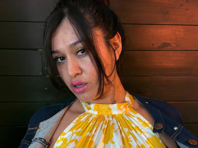 AbbyMontgomery with dark hair, pink lipstick, yellow and white patterned halter top, denim jacket, tattoo on neck, looking slightly bored, wooden wall in background.