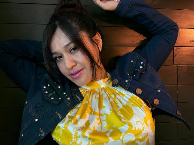 AbbyMontgomery with yellow and white floral top, denim jacket, arms above head, dark hair in bun, pink lipstick, smiling, wooden background, chest visible, no visible genitals.