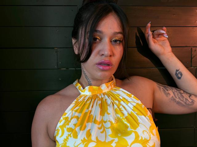 AbbyMontgomery with yellow and white floral halter top, pink lipstick, tattoos on neck and arm, dark hair in ponytail, leaning against dark wood wall, hand up.