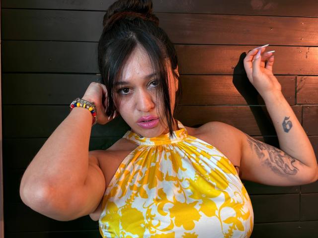 AbbyMontgomery with dark hair in a bun, wearing a yellow floral halter top, has pink lipstick, a tattoo on her right arm, and colorful bracelets, standing against a dark wall.