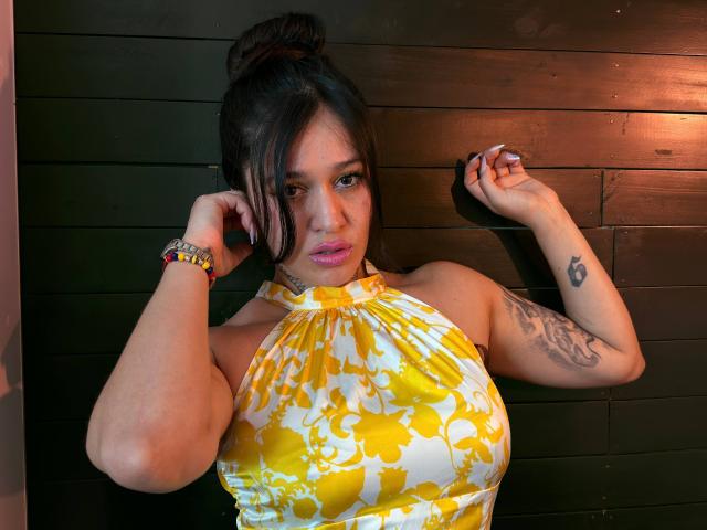 AbbyMontgomery with yellow floral halter top, pink lipstick, black hair in bun, tattoos on right arm, bracelet, leaning against dark wooden wall, hands up, sexy, curvy, tits, hot.