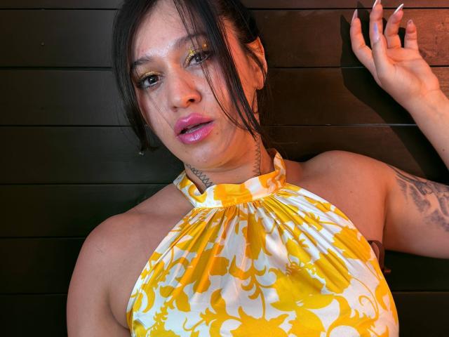 AbbyMontgomery with yellow and white floral halter top, glossy pink lips, dark hair, gold eye makeup, tattoo on neck, black wood background, and raised right hand.