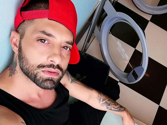 TommyDum, with a red cap, black shirt, and tattoos, leans against a wall, sporting a beard and tattoos on his neck and arm. Checkered floor, ring light.
