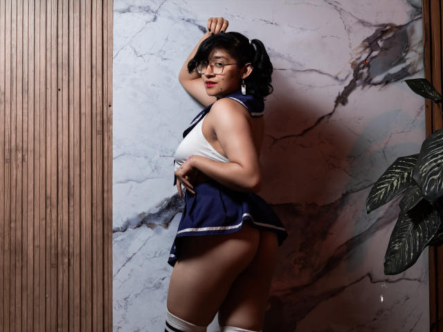 DaryMoon with puffy ass in a sailor-style outfit, white tank top, navy skirt, thigh-high socks, glasses, and black pigtails, standing against a marble wall, arm raised.