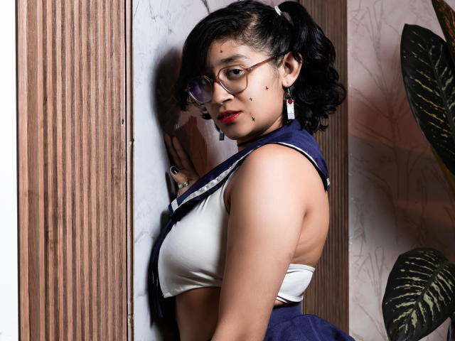 DaryMoon with puffy black hair in pigtails, wearing glasses, red lipstick, and a navy-blue and white schoolgirl outfit, showing her tits, against a wooden wall.