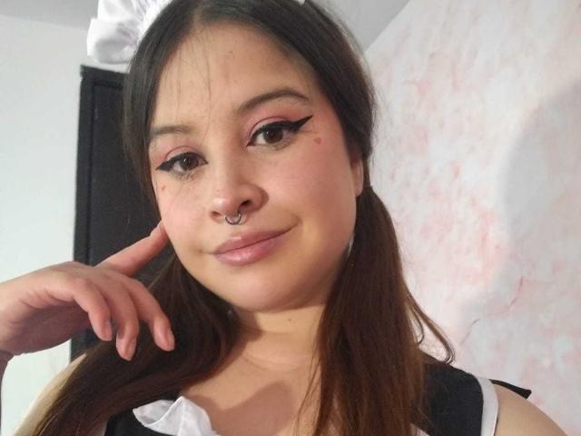 MelaniBecker has brown pigtails, pink eyeliner, and a nose ring. She's smiling with her finger on her cheek, showing her puffy lips and a small red blemish.