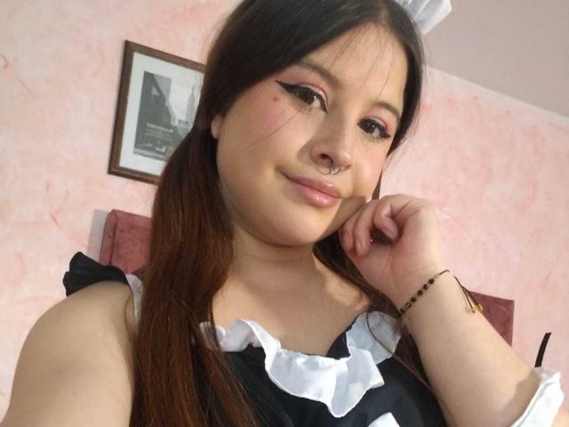 Melani Becker, avec des couettes, un anneau au nez, un eye-liner rose et une tenue de femme de ménage noir et blanc, souriante, la main près du visage, un mur rose, une photo encadrée et une tête de lit rose en arrière-plan..