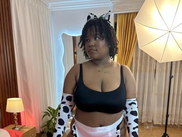 YarhaValois, com pele escura, cabelo cacheado, usando um chapéu com estampa de vaca, um top de ginástica preto e mangas com estampa de vaca, exibindo seus seios grandes e uma calcinha branca pequena..