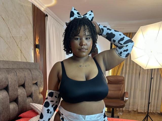 YarhaValois, con piel oscura, cabello rizado, vistiendo un top deportivo negro, mangas con estampado de vaca y una diadema con estampado de vaca, de pie en una habitación con una cama y luces de estudio..