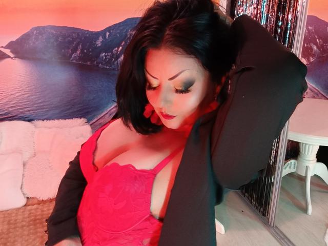 AmberSunshine with big tits in a red lace top, black jacket, and red earrings, looking down with her hand in her hair, in a colorful, warm-lit room.
