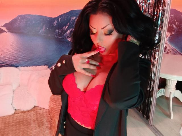 AmberSunshine with black hair, red lace top, and black blazer, touching her red-lipped, cleavage-focused face, finger on her lower lip, showing off her big tits.