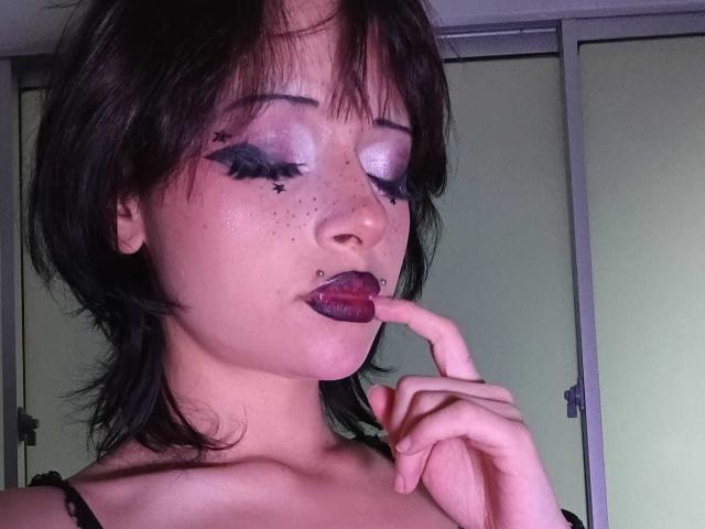 ViolettClay met donker haar, paarse oogschaduw, sterren eyeliner, sproeten, zwarte lipstick, vinger in de mond, tong uit, zwarte bandjes, bleke huid, gemiddelde borsten, slaapkamer, zacht licht..