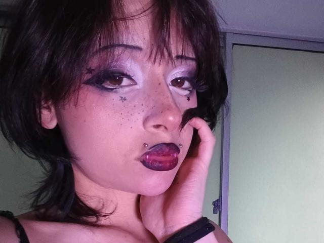ViolettClay has dark brown hair, star freckles on her cheeks, purple lipstick with a lip piercing, and is touching her face with a hand. She's wearing black eyeliner.