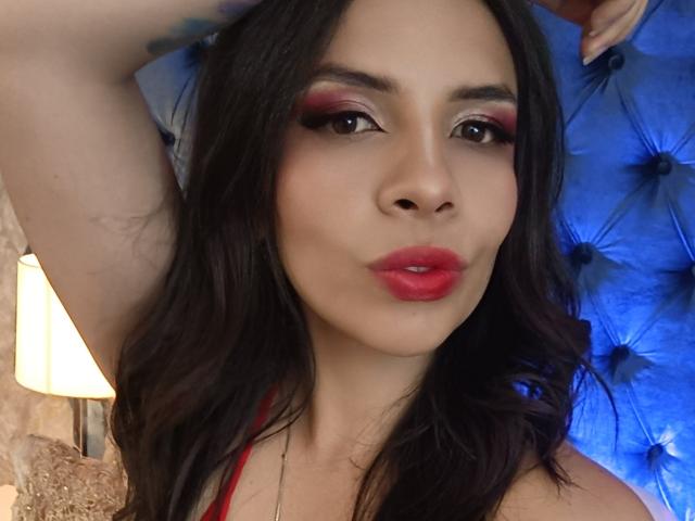 VioletaStone has red lipstick, dark wavy hair, and smoky eye makeup, with one arm raised, showing a tattoo. Her tits are slightly visible in a red top.