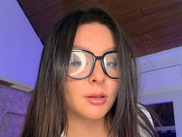 EmaJames, avec ses cheveux bruns lisses, porte des lunettes noires, a une peau pâle, des lèvres roses, et regarde légèrement sur le côté avec une expression neutre..