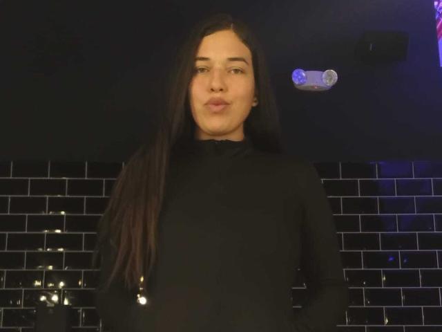 Rachel Wilson, con lunghi capelli castano scuro, un maglione nero a collo alto e labbra piene, in piedi in una luce fioca, che guarda la fotocamera, con le braccia lungo i fianchi, e uno sfondo con una parete di piastrelle nere..