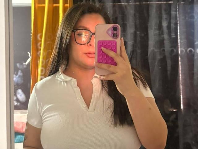 EmaJames, avec de longs cheveux foncés, des lunettes, une chemise blanche côtelée, tenant un téléphone rose, prenant un selfie devant un miroir, montrant ses seins, lèvres roses et douces, rideau noir en arrière-plan..