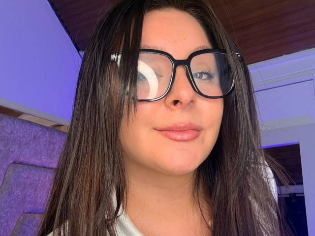 EmaJames, avec de longs cheveux bruns, des lunettes noires et du rouge à lèvres rose, affichant un léger sourire. Son visage est centré, et sa chemise blanche est à peine visible..