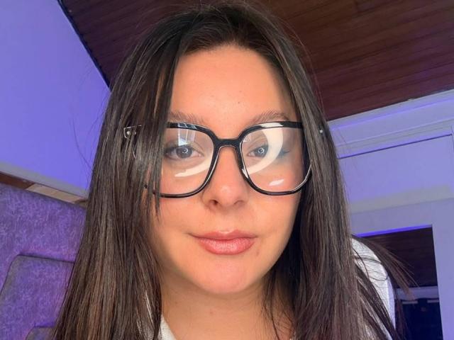 EmaJames, avec des lunettes, des cheveux bruns lisses et une peau claire, a un sourire discret et une petite imperfection sur sa joue droite, et elle porte une chemise blanche..