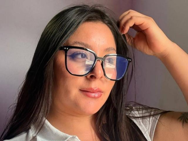 EmaJames, avec ses lunettes noires, ses cheveux bruns brillants et sa chemise blanche, touche ses cheveux, reflétant ainsi le bleu de son œil dans le verre. Ses seins sont discrets..