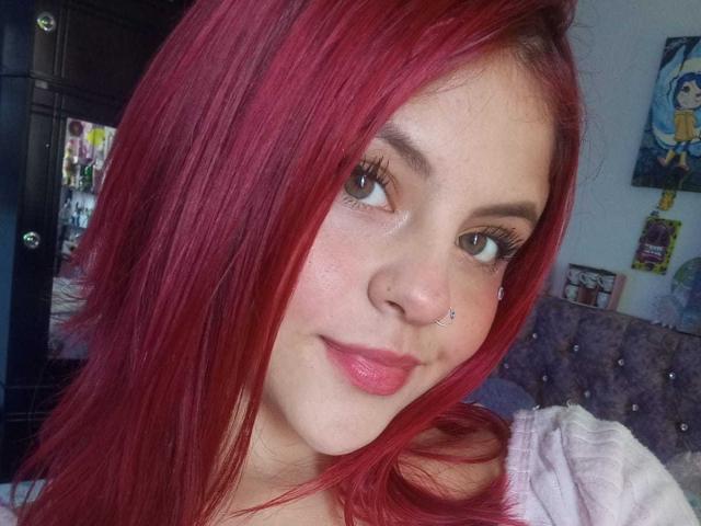 SophieHill has red hair, brown eyes, and a small nose piercing with a septum ring. She's smiling with pink lips, wearing a light pink top.