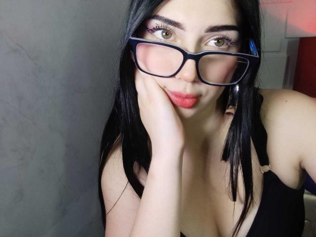IsabellaaRossi with black glasses, red lipstick, and hand on cheek, wearing a black tank top, showing her tits and looking cute with brown eyes and straight black hair.