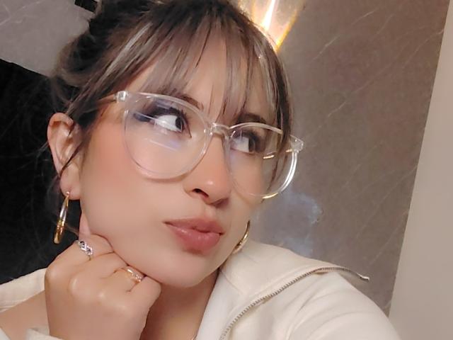 AmiiRosse con gli occhiali, labbra tumefatte e una mano sul viso, indossa una felpa bianca, orecchini a cerchio dorati e un anello, e guarda di lato. I suoi capelli castani sono raccolti in uno chignon..