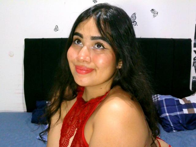 SweetieEma mit langen, schwarzen Haaren, einem roten Spitzen-Haltertop, das ihre nackten Schultern zeigt, und rotem Lippenstift, lächelt in die Kamera, ihre braunen Augen schauen leicht nach oben..