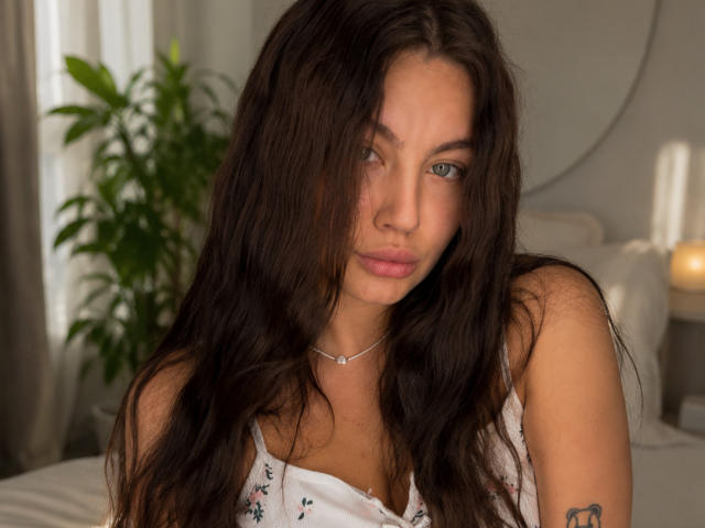 Ines Gueningsman, com cabelos castanhos longos, olhos azuis e lábios rosados, vestindo uma blusa branca com estampas florais, um colar pequeno e uma tatuagem no braço direito. Ela está olhando para a câmera..